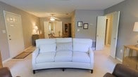 This single-floor residence offers a thoughtfully arranged layout that blends comfort and functionality in a warm, inviting atmosphere. The open-concept design connects the living room and dining area seamlessly, creating a generous sense of flow and space. The living room is anchored by a gently curved, light-colored sofa that adds a touch of elegance to the relaxed setting, complemented by soft carpeted flooring and neutral gray and beige wall tones that lend a calm, cohesive feel throughout. A ceiling fan overhead provides both light and airflow, enhancing the room's everyday comfort. Framed artwork on the walls adds a personal touch to the space. The adjacent dining area is furnished with a wooden table and chairs, accompanied by a bookcase or display cabinet that adds warmth and character. A mirror near the dining space reflects light and contributes to a sense of openness. Interior doors provide access to additional rooms, suggesting a well-organized floor plan with private spaces tucked conveniently beyond the main living areas. Overall, this home presents a tidy, move-in-ready interior with a classic residential aesthetic that prioritizes comfort and livability.