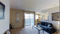 Located at 2677 Collinford Dr in Dublin, Ohio, this single-floor residence offers a well-connected living and dining layout designed for comfortable everyday living. The living area is anchored by plush wall-to-wall carpeting and a large sliding glass door dressed with vertical blinds and decorative curtains, flooding the space with natural light while providing direct access to an outdoor patio. A full-length wall mirror adds depth and dimension to the room, while the open flow into the dining area creates a seamless, sociable atmosphere. The dining space is illuminated by an elegant chandelier and is positioned near an additional window, enhancing the bright and airy feel throughout. A pass-through or half-wall connects the dining area to the kitchen, promoting easy interaction between spaces. The textured ceilings add a classic touch throughout the home. Overall, the space presents a warm, neutral palette with beige tones and thoughtful lighting that contributes to an inviting and functional living environment, ideally situated in the desirable Dublin, Ohio community.