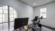 The Spin Room is a purpose-built indoor cycling studio designed to deliver a focused and energizing workout experience. The room is outfitted with multiple Peloton stationary bikes, each equipped with individual touchscreen monitors, allowing users to engage with on-demand and live cycling classes. The bikes are arranged in rows on durable, dark-toned carpet flooring with yellow lane markings that add a structured, studio-like feel to the space.

Architecturally, the room features elegant crown molding and crisp white walls that create a clean, motivating atmosphere. Natural light flows in generously through multiple windows, including a distinctive arched window that adds a refined architectural touch. Additional standard grid-pane windows along the walls further brighten the interior and provide views of the surrounding outdoor greenery.

Access to the spin room is through a sleek, floor-to-ceiling glass sliding door with a matte black handle, offering a modern and transparent boundary between the studio and the adjacent hallway. Recessed ceiling lighting supplements the natural light, ensuring the space remains well-illuminated throughout the day. The overall design balances functionality with aesthetic appeal, making this spin room a well-appointed fitness destination suited for both casual riders and dedicated cycling enthusiasts.