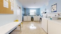 Located at 2243 Eglinton Ave E in Toronto, this shared laundry facility offers a clean, efficiently organized space designed to accommodate multiple users. The room is finished with light blue walls and a white ceiling, creating a bright and airy atmosphere, complemented by a terrazzo floor that is both durable and easy to maintain. A row of top-load washing machines lines one wall, paired with matching front-load washers and dryers along another, providing ample capacity for residents. A deep double utility sink with plumbing fixtures is available for handwashing or pre-treating garments, positioned alongside white subway tile walling for easy cleanup. A centrally placed white folding table and yellow chairs offer a practical space for sorting and folding laundry. Natural light enters through a window along the back wall, enhancing the room's overall brightness. The space is equipped with a wall-mounted fire extinguisher, a hand sanitizer dispenser, informational bulletin boards with posted instructions and notices, and a wall-mounted HVAC unit for climate control. The facility is accessible via a single entry door, making it a straightforward and functional amenity for building residents.