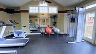 This fitness center offers a comprehensive workout environment designed to accommodate a wide range of exercise routines. The space features soaring vaulted ceilings accented by a warm two-tone color scheme of cream and deep burgundy, creating an energizing yet welcoming atmosphere. A large arched window positioned high on the wall floods the room with natural light, complemented by a ceiling fan that promotes air circulation throughout the space. The rubber-floored workout area is well-appointed with cardio equipment including treadmills and a stationary bike, alongside a multi-station cable strength training machine. A collection of stability and exercise balls in varying sizes adds versatility for core and balance training. Full-length mirrors line one wall, enhancing the sense of openness while allowing for form checks during exercise. Brick-accented double doors provide an attractive entryway, adding a classic architectural touch. The overall layout is thoughtfully organized, offering ample room to move freely between stations, making it suitable for both individual training sessions and small group workouts.