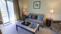 This single-floor space showcases a thoughtfully designed living room that balances comfort and contemporary style. The room is finished with neutral wall tones that create a calm, welcoming ambiance. A plush, gray sofa serves as the centerpiece, accented with coordinating blue and patterned throw pillows that add visual interest. A rectangular coffee table with a light stone-like surface sits atop a soft area rug, styled with decorative trays, candles, and stacked books, adding a lived-in, curated feel. A tall, slender table lamp provides warm ambient lighting, complementing the natural light that filters in through floor-length, textured drape panels framing a window on the left side of the room. A framed abstract artwork adorns the wall above the sofa, contributing to the polished aesthetic. Subtle greenery in the form of small potted plants adds a natural touch to the space. The overall design reflects a cohesive, modern aesthetic with soft furnishings and carefully chosen décor creating an inviting and functional living environment.