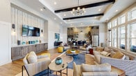 This single-floor community amenity space showcases a thoughtfully designed open-concept layout that seamlessly blends multiple functional zones into one cohesive environment. The main lounge area commands attention with its soaring tray ceiling accented by bold dark wood beams, white shiplap detailing, and a striking sputnik chandelier that anchors the space. Warm hardwood-style flooring runs throughout, complementing the neutral and earthy color palette punctuated by teal and gold accents.

Floor-to-ceiling windows along one wall flood the lounge with abundant natural light and provide views of the outdoor pool area, creating a bright and inviting atmosphere. Multiple seating arrangements — including plush sofas, woven accent chairs, and vibrant yellow ottomans — offer flexible gathering options suited for both casual lounging and social interaction.

A built-in kitchenette at the rear of the space features rich wood cabinetry with glass-front uppers, open shelving with decorative accents, a subway tile backsplash, and a full-size stainless steel refrigerator, making it well-equipped for entertaining. Adjacent dining areas with dark-framed chairs and round tables provide additional seating for meals or meetings.

A semi-private room with a round dining table, teal upholstered chairs, and a bold abstract art piece offers a quieter setting for smaller gatherings, with direct access to the outdoor patio area.