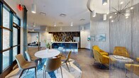 This expansive single-floor amenity space is thoughtfully designed to offer a variety of social and recreational experiences within a cohesive, contemporary aesthetic. The open-plan layout flows seamlessly between distinct zones, each characterized by carefully curated furnishings and architectural details.

A central lounge area features a striking cylindrical chain-curtain enclosure suspended from a circular ceiling detail, housing mustard-yellow velvet armchairs around a low table — an intimate seating nook within the larger space. Nearby, a round marble-top dining table paired with leather chairs sits atop a textured area rug. Large-format hexagonal floor tiles in contrasting tones define the bar area, which features a generous white quartz-topped island with built-in television screens and upholstered bar stools.

A dedicated golf simulator room with a large projection screen and carpeted floor provides an immersive recreational experience, while a ping-pong table anchors a games zone with direct views to an outdoor area through expansive glass doors. A cozy lounge nook with a sofa, accent chairs, and vibrant abstract artwork offers a quieter retreat. Throughout, polished concrete floors, pendant lighting, bold accent walls, and colorful art installations create a dynamic, welcoming ambiance.