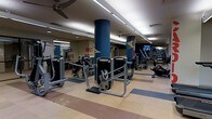 This fitness center is designed to accommodate a comprehensive workout experience within a single-level, open-plan layout. The space is anchored by bold navy blue cylindrical columns that add a structural yet design-forward element throughout the floor. Brightly colored motivational text and graphic accents adorn the walls, contributing to an energetic atmosphere. The flooring features a multi-toned tile pattern in earth and cool tones, delineating workout zones while adding visual interest. Overhead, a combination of recessed lighting and linear fluorescent fixtures ensures thorough illumination across the entire space, supplemented by ceiling fans that aid in air circulation. A mirrored wall on one side visually expands the area and allows users to monitor their form. The equipment layout is well-organized, with strength-training machines positioned centrally and cardio equipment, including rowing machines and stationary bikes, occupying the perimeter. A mounted flat-screen television is visible toward the rear, offering entertainment during workouts. The overall ambiance is functional, bright, and motivating.