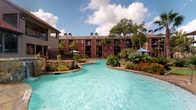 This pool clubhouse facility offers an inviting combination of indoor comfort and outdoor recreation. At the heart of the outdoor area is a sprawling, freeform lagoon-style pool with crystal-clear turquoise water, framed by lush tropical landscaping, decorative stonework, and a spacious deck. Covered canopy areas line the poolside, providing shaded gathering spots, while wrought iron fencing and mature trees enhance the resort-like atmosphere surrounding the multi-story brick residential buildings.

Inside, the clubhouse is anchored by a bright, open-plan interior with tall ceilings, crown molding, and large-format tile flooring throughout. The main lounge area benefits from generous natural light streaming through ornate glass-paneled entry doors, complemented by a ceiling fan and elegant chandelier. A seating area with wicker chairs adds a casual, welcoming touch.

Adjacent to the lounge is a well-appointed community kitchen featuring rich dark wood cabinetry, granite countertops, stainless steel appliances including a refrigerator and microwave, and a central island with bar seating. High-top tables and chairs provide additional dining and socializing space. Large windows offer direct views of the pool deck, seamlessly connecting the indoor and outdoor environments and reinforcing the space's vibrant, community-focused character.