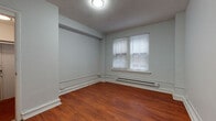 This single-floor space is characterized by a cohesive design that balances functional living areas with updated finishes throughout. Rich hardwood flooring in a warm walnut tone flows consistently across the main living and bedroom areas, complementing the soft grey-blue walls and white baseboards that frame each room. Textured ceilings and flush-mount lighting fixtures are present throughout, while baseboard heating units run along the lower walls beneath windows dressed with white horizontal blinds, allowing for controlled natural light.

The bedroom areas are well-proportioned, each offering a open, uncluttered layout with structural beam details visible along the upper walls. Each room benefits from natural light through double-pane windows and provides practical living space suitable for standard furnishings.

The kitchen is outfitted with dark espresso cabinetry, granite countertops, a stainless steel sink, a gas range, and an over-the-range microwave. A white subway tile backsplash adds a clean, contemporary contrast to the darker cabinetry.

The bathroom features a glass-enclosed walk-in shower with white horizontal tile walls, a decorative mosaic tile accent border, a mosaic tile shower floor, and a standard white toilet, creating a clean and modern aesthetic throughout.
