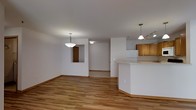 This single-floor space presents an open, flowing layout that connects the main living area to the kitchen and additional rooms beyond. Warm-toned hardwood flooring extends throughout the primary living space, lending a cohesive and inviting feel to the interior.

The kitchen occupies the right side of the space and is defined by a curved breakfast bar counter that serves as a natural divider between the kitchen and living area. Medium-toned wood cabinetry lines the upper kitchen walls, complemented by a white refrigerator and track lighting overhead. A pendant light hangs above the bar area, adding focused illumination.

The living area is generously proportioned, with neutral white walls and smooth ceilings. A bowl-style pendant light fixture anchors the dining or living zone, providing ambient light. A pass-through window or recessed nook is visible along one wall, adding architectural interest.

Beyond the main living space, a hallway leads to additional rooms, where the flooring transitions to carpet. The overall palette is clean and neutral, with white walls and natural wood tones creating a bright, understated ambiance throughout the space.