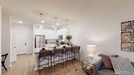 This single-story residence offers a thoughtfully arranged open-concept layout that seamlessly connects the main living spaces. The kitchen is a standout feature, outfitted with crisp white cabinetry, granite-style countertops, and a full suite of stainless steel appliances, including a refrigerator, electric range, and microwave. An extended breakfast bar with seating for several provides a practical and social dining option, illuminated overhead by elegant pendant lighting. Light wood-style flooring flows throughout the main living areas, enhancing the bright, airy atmosphere.

The living room is a comfortable and well-lit space, anchored by natural light and access to a private balcony through full-length French doors dressed with linen-style curtains — an inviting outdoor extension of the interior.

The bedroom is a calm and restful retreat with carpeted flooring, generous wall space, and access to a dedicated walk-in closet equipped with wire shelving and hanging rods. The full bathroom connects conveniently to the bedroom and features granite-topped vanity cabinetry, a large mirror, and a combination tub and shower. An in-unit washer is also visible, adding everyday practicality to this well-appointed residence.