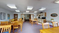 This single-floor communal space is thoughtfully designed to serve as a shared dining and gathering area. The main room is generously sized, accommodating multiple rectangular dining tables paired with light wood chairs, arranged to encourage both group dining and social interaction. The space is bathed in bright, even light provided by a series of large, frosted panel overhead light fixtures that lend a clean, airy feel to the interior.

The flooring features soft purple carpeting throughout the dining area, adding warmth and comfort underfoot. Along one side of the room, a functional kitchenette is integrated seamlessly into the layout, featuring upper cabinetry, countertop workspace, a sink, a refrigerator, and small appliances including a coffee maker and microwave — making it well-suited to support meal service and refreshments.

French-style glass-paned doors lead to adjacent rooms, suggesting additional gathering or activity spaces beyond the main area. Touches of greenery, including potted plants and small floral arrangements on the tables, add a welcoming, homey atmosphere. The neutral walls and practical layout reflect a design intended to comfortably serve a community of residents or visitors in a functional yet inviting setting.
