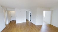 This single-floor space showcases a clean, open-concept layout designed to maximize functionality and flow. The main living area features warm-toned hardwood flooring that extends throughout, complementing the bright white walls and creating a welcoming atmosphere. The layout is defined by partial walls that divide the space into distinct zones while maintaining an overall sense of openness.

Toward the back, a passageway leads to the kitchen area, where a stainless steel refrigerator is visible, hinting at modern appliances within. The kitchen appears to be a separate, enclosed space accessible from the main living area. A window is visible to the right side of the space, allowing natural light to filter in and brighten the interior. The flooring transitions near this area, suggesting a defined secondary zone, possibly a dining or additional living space.

The white walls are smooth and neutral, providing a versatile backdrop suitable for a variety of uses. The layout is efficient, with clear sightlines from one end to the other. Overall, the space presents a straightforward, functional design with an airy, minimalist character.