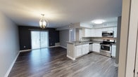 This single-floor home offers three bedrooms and two bathrooms within a well-designed open-concept layout. The kitchen is a standout feature, outfitted with crisp white cabinetry, granite countertops, and stainless steel appliances including a range, microwave, and dishwasher. A peninsula with a sink extends into the living area, creating a natural gathering space and enhancing the flow between the kitchen and main living areas. Dark hardwood-style flooring runs throughout, adding warmth and visual continuity across the space. The living and dining areas are defined by a bold navy accent wall and a decorative globe chandelier that adds a contemporary touch. A pendant light over the peninsula echoes this design detail. Sliding glass doors in the living area fill the space with natural light and provide access to the outdoors. The overall aesthetic is modern and clean, combining neutral gray tones with contrasting dark flooring and bold accent colors for a polished, cohesive look.