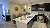 This thoughtfully designed single-floor residence showcases a seamless open-concept layout that connects the kitchen, dining, and living areas into one cohesive and functional space. The kitchen is a standout feature, outfitted with rich dark cabinetry that contrasts beautifully against crisp white countertops. A generously sized island with bar seating serves as both a casual dining spot and a central gathering point, complete with a stainless steel undermount sink and elegant pendant lighting overhead. Stainless steel appliances, including a built-in microwave, range, and dishwasher, add a polished, contemporary feel, while a classic tile backsplash provides a refined finishing touch. Light-toned hardwood-style flooring runs throughout, lending warmth and continuity across the open floor plan. The dining area flows naturally from the kitchen, offering an intimate setting for meals and entertaining. Soft, neutral wall tones create a calming backdrop that complements the modern furnishings and decorative wall art. Recessed lighting combined with stylish pendant fixtures ensures the space is well-lit and inviting, making this a comfortable and visually appealing home.