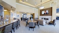 This elegantly appointed single-floor communal space exudes a refined, upscale ambiance designed for both relaxation and social gatherings. The room immediately impresses with its soaring tray ceilings, accented by recessed cove lighting that casts a warm, inviting glow throughout the space. A large pendant light anchors the central seating area, complementing the carefully layered lighting scheme that includes track lighting and wall sconces.

The layout is thoughtfully organized into distinct zones. A lounge area features plush, neutral-toned sofas and armchairs arranged around a sleek fireplace with a stone surround, above which a flat-screen television is mounted — creating a cozy, living room-like atmosphere. Adjacent to this, multiple round dining tables with upholstered chairs in a contemporary patterned fabric provide ample seating for gatherings or casual dining.

The warm beige walls are adorned with framed artwork, adding a polished, gallery-like touch. Large windows and glass doors along one wall allow natural light to filter in while offering views of the exterior, enhancing the open and airy feel of the space. Lush potted plants are strategically placed throughout, introducing organic warmth and color. The plush carpeted flooring ties the entire space together with a sense of comfort and sophistication.