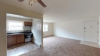 Located at 835 Blooming Grove Turnpike in New Windsor, NY, this single-floor space offers a straightforward and functional layout designed for comfortable everyday living. The open-concept design connects the kitchen and living area, creating a cohesive flow throughout the unit. The kitchen is equipped with stainless steel appliances, including a gas range, and features warm wood-toned cabinetry with under-cabinet ventilation. Checkered tile flooring in the kitchen provides a classic, easy-to-maintain surface. The living area transitions seamlessly from the kitchen via an open pass-through wall, and is finished with plush wall-to-wall carpeting that adds warmth underfoot. A ceiling fan with a wood-toned blade accent overhead provides both air circulation and a subtle decorative touch. Natural light filters into the living area through a window, brightening the neutral, cream-toned walls. Baseboard heating runs along the perimeter, ensuring consistent warmth throughout the cooler months. The overall ambiance is clean, neutral, and move-in ready, offering a practical living environment in a convenient New Windsor location.