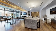 This contemporary amenity lounge is thoughtfully designed as a multi-functional social space, seamlessly blending comfort, style, and entertainment across a single open floor plan. Warm hardwood flooring runs throughout, anchoring the space with an inviting, cohesive aesthetic.

At the heart of the lounge sits a striking marble island with substantial seating, ideal for gathering and conversation. Nearby, a fully equipped wet bar features stainless steel appliances, under-counter refrigeration, a wall-mounted TV, and clean white cabinetry — providing everything needed for entertaining. A decorative slatted wood ceiling element with integrated lighting adds architectural interest overhead, while a modern branching chandelier illuminates the central area with ambient warmth.

The lounge area offers plush seating arranged around a sleek linear fireplace set against a dark tile surround, accompanied by a large wall-mounted television and floor-to-ceiling glass walls that flood the space with natural light and open directly to an outdoor terrace. A textured, upholstered banquette nook with padded wall panels provides an intimate retreat for smaller gatherings.

The game zone features an air hockey table, wall-mounted display, and bar-height seating along a built-in ledge, catering to recreational use. Lush potted greenery is distributed throughout, softening the modern design and adding a natural, refreshing element to the overall ambiance.