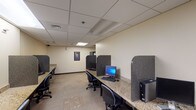 This business center is a well-organized, single-floor facility designed to support productivity and convenience. The space is laid out with two parallel runs of granite-topped counters lining the walls, each accommodating multiple individual workstations equipped with desktop computers, monitors, keyboards, and mice. Dark granite privacy dividers separate each station, providing users with a degree of personal space while maintaining an open, communal atmosphere.

The room features a drop ceiling with recessed panel lighting and skylight-style flush fixtures that provide consistent, even illumination throughout. Neutral beige walls and light-toned carpet flooring contribute to a clean and professional environment. A separate alcove area adjacent to the main workstation room houses a vending machine alongside a small seating area with a compact chair, offering a convenient spot for short breaks.

An additional dedicated workspace is available in the form of a private U-shaped granite desk area, partitioned from the main room, providing a more enclosed setting for focused tasks. Decorative framed artwork accents the walls, adding a subtle visual element to the otherwise utilitarian space. The overall layout is efficient and practical, making the business center a well-equipped resource for those needing computer access or a quiet place to work.