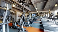 The fitness center at Baypointe is a generously proportioned, single-floor facility designed to support a comprehensive workout experience. The space features an industrial-inspired aesthetic, with an exposed ceiling showcasing structural beams, metal ductwork, and a combination of large ceiling fans and warm pendant lighting that together create an energetic yet comfortable atmosphere.

The cardio zone is a standout feature, lined with rows of elliptical trainers and treadmills arranged in an orderly fashion along rubberized flooring accented with orange striping. Mounted televisions are positioned throughout the space, allowing members to stay entertained during workouts. Strength and resistance training equipment is also present, rounding out the facility's offerings for a full-body training experience.

Large wall mirrors extend along one side of the gym, visually expanding the space and providing functional feedback during exercise. The combination of durable rubber flooring, ample equipment, and thoughtful lighting makes this fitness center a well-appointed amenity that balances functionality with a modern, urban design sensibility.