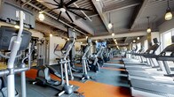 The fitness center at Baypointe is a generously proportioned, single-floor facility designed to support a comprehensive workout experience. The space features an industrial-inspired aesthetic, with an exposed ceiling showcasing structural beams, metal ductwork, and a combination of large ceiling fans and warm pendant lighting that together create an energetic yet comfortable atmosphere.

The cardio zone is a standout feature, lined with rows of elliptical trainers and treadmills arranged in an orderly fashion along rubberized flooring accented with orange striping. Mounted televisions are positioned throughout the space, allowing members to stay entertained during workouts. Strength and resistance training equipment is also present, rounding out the facility's offerings for a full-body training experience.

Large wall mirrors extend along one side of the gym, visually expanding the space and providing functional feedback during exercise. The combination of durable rubber flooring, ample equipment, and thoughtful lighting makes this fitness center a well-appointed amenity that balances functionality with a modern, urban design sensibility.