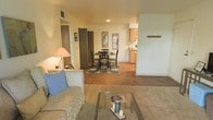 This single-story residence showcases an open-concept floor plan that seamlessly connects the living, dining, and kitchen areas, creating a cohesive and functional living environment. The living area is carpeted and anchored by a plush sofa with accent pillows and a glass-top coffee table, lending a relaxed, welcoming atmosphere. Warm-toned hardwood flooring extends through the dining and kitchen zones, providing a natural visual transition between spaces. The dining area accommodates a round table with seating for four, positioned atop a patterned area rug and framed by wall art that adds a decorative touch. Beyond the dining space, the kitchen features light wood cabinetry and appears well-equipped for everyday cooking. A coat hook rail is mounted near what appears to be an entryway, adding a practical organizational element. Neutral wall tones and recessed lighting throughout contribute to a bright, airy feel. A doorway near the living area suggests access to additional private spaces such as a bedroom or bathroom, completing the layout of this well-appointed single-floor residence.
