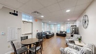 This single-floor multipurpose space is thoughtfully designed to serve as a welcoming communal gathering area. The open lounge area is the heart of the space, featuring warm hardwood flooring, a drop ceiling with recessed lighting, and multiple windows dressed with horizontal blinds that invite abundant natural light and views of the surrounding outdoor greenery. The lounge is furnished with dark leather sofas and loveseats, accent chairs with bold patterned upholstery, and a media console with a flat-screen television, creating a relaxed and functional environment. A round dining table with chairs near the entrance provides a dedicated spot for small group meetings or casual meals. The kitchenette is well-appointed with warm wood cabinetry, granite-style countertops, a stainless steel sink, and a full-size refrigerator, offering practical amenities for gatherings or events. A dedicated ADA-accessible restroom features elegant marble-style tile flooring, stainless steel grab bars, and a frosted privacy window, ensuring both accessibility and comfort. Throughout the space, neutral wall tones and consistent flooring tie each area together into a cohesive, functional, and inviting environment suitable for a variety of community or organizational uses.