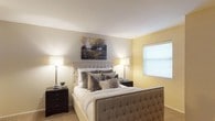 This single-story space showcases a thoughtfully designed bedroom that combines comfort and style. The room is finished with soft yellow walls that lend a warm, welcoming ambiance, complemented by white ceilings that help brighten the interior. Natural light filters in through a window fitted with white blinds, adding to the airy feel of the space.

The bedroom centers around a generously sized upholstered bed with a tufted headboard and footboard in a neutral taupe tone, accented with nailhead trim detailing that adds a refined touch. The bed is dressed in crisp white linens layered with an assortment of textured throw pillows in grey and silver tones, contributing to the room's cohesive, elegant aesthetic.

Flanking the bed are two dark wood nightstands, each topped with a matching slim chrome table lamp that provides soft, ambient lighting. A landscape canvas artwork hangs above the headboard, depicting trees in muted earth tones that complement the room's color palette. Plush wall-to-wall carpeting covers the floor, enhancing the overall sense of warmth and comfort throughout the room.