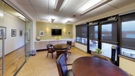 This single-floor professional gathering space is designed to facilitate comfortable, collaborative interaction in a polished environment. The room features cork flooring throughout, lending a warm, natural texture underfoot, complemented by a suspended tile ceiling with recessed and fluorescent lighting that ensures consistent, well-distributed illumination.

Two round dark-wood tables, each surrounded by leather-upholstered chairs on casters, serve as the primary seating arrangements, supporting both formal and informal meetings. Along one wall, a long white built-in credenza provides generous storage beneath a wall-mounted flat-screen television, flanked by decorative sconce lighting and framed artwork. A globe and floral accents contribute a refined, welcoming ambiance.

Framed portraits displayed on the sage-green accent walls lend the space a distinguished character. Folding chairs stored along the wall offer flexible capacity for larger gatherings. Glass-panel partitions and doors on one side provide visual connectivity to an adjacent corridor, while large windows fitted with roller shades face an outdoor terrace, offering views and access to the exterior. The overall design balances a professional, institutional aesthetic with approachable warmth, making it well-suited for meetings, social gatherings, or multipurpose community use.