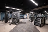 This modern fitness center is thoughtfully designed to accommodate a wide variety of workout routines. The space features a dark, industrial-style ceiling with sleek linear LED lighting that illuminates the floor throughout. Durable rubber and stone-tile flooring provides a sturdy, practical surface suited for high-intensity activity.

The cardio zone is lined along a teal accent wall and includes multiple treadmills, stationary cycling bikes, and an elliptical machine, each equipped with individual display screens for an interactive workout experience. Adjacent to this area, a dedicated strength training section houses a full cable machine system, including a Life Fitness multi-functional unit and a Synrgy360 functional trainer, offering versatile resistance training options.

Free weight equipment is well-represented, with barbells, weight plates, plate racks, and a weight bench available for compound and isolation exercises. A battle rope and stability balls in multiple sizes round out the functional fitness offerings. Heavy punching bags are suspended from the ceiling, adding a boxing and combat training element to the space.

Floor-to-ceiling mirrors line one wall, enhancing the sense of openness and allowing users to monitor their form. The overall ambiance is clean, professional, and well-organized, reflecting a facility designed to meet diverse fitness needs.
