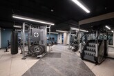 This modern fitness center is thoughtfully designed to accommodate a wide variety of workout routines. The space features a dark, industrial-style ceiling with sleek linear LED lighting that illuminates the floor throughout. Durable rubber and stone-tile flooring provides a sturdy, practical surface suited for high-intensity activity.

The cardio zone is lined along a teal accent wall and includes multiple treadmills, stationary cycling bikes, and an elliptical machine, each equipped with individual display screens for an interactive workout experience. Adjacent to this area, a dedicated strength training section houses a full cable machine system, including a Life Fitness multi-functional unit and a Synrgy360 functional trainer, offering versatile resistance training options.

Free weight equipment is well-represented, with barbells, weight plates, plate racks, and a weight bench available for compound and isolation exercises. A battle rope and stability balls in multiple sizes round out the functional fitness offerings. Heavy punching bags are suspended from the ceiling, adding a boxing and combat training element to the space.

Floor-to-ceiling mirrors line one wall, enhancing the sense of openness and allowing users to monitor their form. The overall ambiance is clean, professional, and well-organized, reflecting a facility designed to meet diverse fitness needs.
