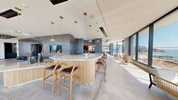 Located at 39 Bruton Street on Yerba Buena Island in San Francisco, this expansive single-floor amenity lounge is a masterfully designed communal space that blends contemporary architecture with refined, hospitality-inspired interiors. Polished concrete floors stretch throughout, reflecting abundant natural light that pours in through dramatic floor-to-ceiling glass walls offering sweeping panoramic views of the San Francisco Bay and surrounding cityscape.

At the heart of the space sits a sculptural oval bar island topped with elegant marble and wrapped in fluted natural wood, surrounded by leather-cushioned bar stools. A striking accent wall clad in vertically stacked blue-grey tile serves as a bold architectural focal point, complemented by a mounted flat-screen display. Brass pendant lights hang in a cascading cluster above the bar, adding warmth and a refined touch.

A dedicated back bar area features moody oxidized metal surfaces, mirrored ceilings with geometric brass accent lighting, light wood cabinetry, and full bar service provisions. Throughout the lounge, thoughtfully arranged seating zones include a cozy built-in banquette with slatted dark wood paneling, casual bar-height nooks, and an open lounge area with a curved leather sofa and organic-shaped coffee table. The space is completed by an elevator lobby clad in rippled metallic tile, offering a polished arrival experience.