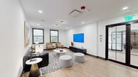 Located at 285 Uptown Blvd in Altamonte Springs, Florida, this thoughtfully designed single-floor amenity space offers a blend of collaborative and focused work environments across three distinct areas. The main lounge serves as the centerpiece, featuring light wood-look flooring, crisp white walls, and a bold geometric cross-shaped pendant light fixture. A sleek black leather sofa, tan leather accent chairs, and rounded upholstered ottomans are arranged atop a graphic patterned area rug, creating a welcoming and versatile gathering area. A wall-mounted flat-screen display and a low media console anchor one wall, while large black-framed windows invite natural light and views of the surrounding Uptown district. A glass-paneled partition with black steel framing provides visual separation while maintaining an open feel, leading to a semi-private lounge nook accented by a dramatic black lattice-screen divider and warm caramel leather booth seating. The dedicated conference room features a rich dark oval table surrounded by tan leather chairs, stylish wall art, a multi-arm chandelier, and natural light through black-framed windows. A compact workstation with bar-height seating rounds out the space, offering additional focused work options. Throughout, the design maintains a cohesive modern aesthetic with carefully curated artwork and finishes.