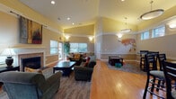 This single-floor community space is designed to accommodate a variety of social and relaxation needs within a warm, welcoming environment. The main lounge area immediately impresses with its vaulted ceilings painted in a soft yellow tone, recessed lighting, and warm hardwood flooring that runs throughout. A classic brick fireplace with a white mantel serves as the room's focal point, flanked by large windows that invite ample natural light and views of the surrounding exterior. Multiple seating arrangements, including plush green sofas and armchairs positioned around area rugs and coffee tables, create distinct zones for gathering or quiet relaxation. Decorative wall art and a painted tree mural add character to the space.

Adjacent to the main lounge, a dedicated seating nook provides additional chairs and a console table, functioning as a quieter retreat. A separate café-style area features a granite-topped bar counter with high stools and clerestory windows positioned near the ceiling, allowing natural light while maintaining privacy. This zone appears suited for casual dining or refreshments.

Throughout the space, wall sconces, pendant lights, and recessed fixtures ensure consistent, comfortable illumination. The overall ambiance is inviting and community-oriented, balancing functional furnishings with tasteful decorative touches.