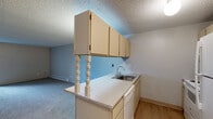 Located at 401 E Magnesium Rd in Spokane, WA, this one-bedroom, one-bathroom unit offers a functional single-floor layout with a well-defined mix of living spaces. The combined kitchen and living room forms the heart of the home, where a peninsula countertop with light stone-style laminate surfaces and decorative turned columns separates the two areas. The kitchen is equipped with white appliances including a range and refrigerator, ample cream-toned cabinetry overhead and below, a stainless steel sink, and light hardwood-style flooring. A pendant chandelier adds a touch of character above the peninsula. The living room features carpet flooring, a textured ceiling, vertical wood panel accent wall, and a sliding glass door that opens to a private balcony with outdoor views. Baseboard heaters are present throughout the unit for consistent warmth. The bedroom offers carpet flooring, a window with a view of the surrounding greenery, and a wall-mounted air conditioning unit. The bathroom includes a full tub/shower combo with white tile surround, hardwood-style flooring, and a vanity with matching light countertop. A dedicated closet and hallway complete the thoughtfully organized floor plan.