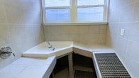 This single-floor space includes a utilitarian wet room or washroom area finished in neutral beige ceramic wall and floor tiles. The room features an L-shaped built-in tiled bench or ledge that wraps along two walls, providing functional seating or a washing surface. A flexible handheld shower hose and fixture is mounted to the countertop ledge, suggesting use as a washing or bathing station. Anti-slip rubber drainage mats are placed on the floor, emphasizing the wet-use nature of the space. Two double-hung windows positioned along the upper wall allow natural light to filter in, brightening the otherwise compact interior. The overall aesthetic is practical and straightforward, with consistent tile work covering both the walls and structural elements throughout the room.
