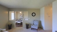 This single-story space presents a straightforward, functional layout characterized by neutral wall tones and a cohesive color palette throughout. The living area features wall-to-wall carpeting underfoot and a textured popcorn ceiling overhead, lending a casual, comfortable atmosphere. A decorative round wall clock serves as a focal point on the main wall, while a patterned accent chair adds a subtle touch of personality to the space.

A pass-through opening connects the living area to the adjacent dining room, creating a sense of visual continuity between the two spaces. The dining room features hard-surface flooring, a pendant light fixture providing warm overhead illumination, and a dining set with metal-framed chairs. A decorative wall panel adds an artistic element to the dining area, while a window allows natural light to filter into the room.

The transition between the carpeted living space and the hard-surface dining area is clearly defined by the step-down entryway. A potted plant near the pass-through adds a natural element to the overall aesthetic. The space maintains a clean, minimalist character with simple baseboards and understated décor throughout.