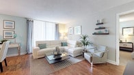This single-floor residence is thoughtfully designed with a clean, coastal aesthetic carried consistently throughout each room. The living area serves as the heart of the home, featuring warm hardwood flooring, a large floor-to-ceiling vertical blind window that draws in generous natural light, and floating wall shelves that add a decorative accent. A generously sized white sectional sofa anchors the space, complemented by a sleek glass-top coffee table and a contemporary accent chair, creating a comfortable and stylish seating arrangement.

Adjacent to the living area, the dining space accommodates a full glass-top dining table with white upholstered chairs, illuminated by a track lighting fixture overhead. A pass-through opening connects the dining room directly to the kitchen, enhancing the open flow of the layout. The kitchen is well-appointed with white cabinetry, granite countertops, a subway tile backsplash, stainless steel appliances, and tile flooring.

The bedroom is a serene retreat, featuring plush carpeting, soft blue accent walls, natural light from a well-positioned window with striped curtains, and a spacious double closet offering ample storage. The overall ambiance of the home is light, airy, and cohesive throughout.