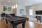 Located at 32 Ashbury Woods Dr in Huntsville, Alabama, this single-floor community clubhouse offers a well-appointed gathering and recreation space designed for both leisure and productivity. The open layout flows seamlessly across warm hardwood flooring and carpeted lounge areas, creating distinct zones for various activities.

At the heart of the space sits a full-size billiards table, illuminated by elegant pendant lights suspended from the ceiling. Adjacent to the pool area, high-backed bar chairs and cue storage provide a dedicated spot for players. A vintage arcade cabinet adds a nostalgic touch near the arched windows, which flood the room with generous natural light and offer views of the surrounding greenery.

The lounge area features a wall-mounted flat-screen TV, a plush area rug, and comfortable accent chairs arranged for relaxed viewing or conversation. Recessed lighting throughout ensures consistent illumination across the entire space.

A dedicated workstation area includes a sleek double-desk setup with ergonomic chairs, ideal for residents needing a quiet place to work or study. Contemporary wall art adds color and personality to the neutral, light-gray walls. Multiple large windows, including arched designs, enhance the airy, open ambiance that defines this versatile community clubhouse.