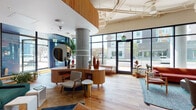 This expansive single-floor communal space blends contemporary architecture with warm, eclectic mid-century modern design. Anchored by prominent exposed concrete columns and wide-plank hardwood flooring, the open floor plan flows seamlessly between several distinct zones, each offering a unique ambiance and function.

The entry area features floor-to-ceiling black-framed glass windows and doors that flood the interior with abundant natural light and provide clear sightlines to the surrounding streetscape. A sculptural, curved wood reception desk with an oversized circular mirror serves as the focal point of the welcoming lobby area.

Multiple lounge zones are thoughtfully appointed throughout the space. A cozy seating nook includes a marble-surround fireplace, wall-mounted display, built-in wood shelving, and a tufted leather sofa. Another area offers a rich arrangement of plaid and leather armchairs, rocking chairs, and lush indoor trees positioned against expansive street-facing windows. Tall wooden bookshelves lined with books and décor add warmth and character.

The kitchen area features stainless steel appliances, subway tile backsplash, a farmhouse sink, and a large island with bar seating. Arched doorways and niches add architectural interest throughout. Pendant lighting, abundant greenery, and vibrant upholstery in emerald, rust, and tan complete the inviting, design-forward atmosphere.