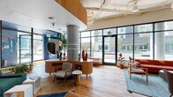This expansive single-floor communal space blends contemporary architecture with warm, eclectic mid-century modern design. Anchored by prominent exposed concrete columns and wide-plank hardwood flooring, the open floor plan flows seamlessly between several distinct zones, each offering a unique ambiance and function.

The entry area features floor-to-ceiling black-framed glass windows and doors that flood the interior with abundant natural light and provide clear sightlines to the surrounding streetscape. A sculptural, curved wood reception desk with an oversized circular mirror serves as the focal point of the welcoming lobby area.

Multiple lounge zones are thoughtfully appointed throughout the space. A cozy seating nook includes a marble-surround fireplace, wall-mounted display, built-in wood shelving, and a tufted leather sofa. Another area offers a rich arrangement of plaid and leather armchairs, rocking chairs, and lush indoor trees positioned against expansive street-facing windows. Tall wooden bookshelves lined with books and décor add warmth and character.

The kitchen area features stainless steel appliances, subway tile backsplash, a farmhouse sink, and a large island with bar seating. Arched doorways and niches add architectural interest throughout. Pendant lighting, abundant greenery, and vibrant upholstery in emerald, rust, and tan complete the inviting, design-forward atmosphere.