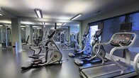 This fitness center is designed to accommodate a comprehensive workout experience within a single-floor layout. The space is outfitted with a variety of cardio equipment, including multiple elliptical trainers and treadmills, positioned to allow comfortable spacing between users. Strength training machines are arranged toward the back of the room, providing a dedicated area for resistance-based workouts. Full-length mirrors line one wall, enhancing the sense of space while allowing users to monitor their form during exercise. The ceiling features industrial-style linear lighting fixtures that provide bright, even illumination throughout the room. The flooring consists of a dark, durable material suited for high-traffic fitness use. Neutral gray walls contribute to a clean, modern aesthetic. A large window on one side of the room allows natural light to enter, adding to the overall ambiance. The layout is thoughtfully organized to maximize usable floor space while maintaining clear pathways between equipment, making the facility both functional and accessible for a range of fitness activities.