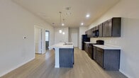 This single-floor residence offers a clean, modern layout designed for comfortable everyday living. The heart of the home is an open-concept kitchen and living area that creates a seamless flow throughout the main space. The kitchen is well-appointed with dark, shaker-style cabinetry extending to the upper walls, light-colored countertops, and a full suite of black appliances including a refrigerator, range, and over-the-range microwave. A generously sized center island provides additional counter space and features a built-in sink, pendant lighting above, and seating overhang — making it ideal for casual dining or entertaining. Light wood-style flooring runs throughout the main living areas, contributing to a bright and cohesive aesthetic. Recessed lighting and a ceiling fan keep the living space comfortable and well-lit. The bedrooms transition to plush carpeting and are finished with neutral-toned walls, white trim, and windows fitted with horizontal blinds that allow for natural light and privacy. The layout, as seen in the dollhouse view, includes multiple bedrooms and at least one bathroom, with a practical and connected floor plan throughout. Overall, the space presents a fresh, move-in-ready environment with a consistent neutral palette and modern finishes.
