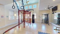 Located at 3001 Bermuda Bay Ln in Mebane, NC, this single-floor fitness center is a comprehensively outfitted facility designed to support a wide range of workout styles. The space is divided into distinct functional zones, each thoughtfully arranged for an optimal training experience.

The main strength and functional training area features soaring double-height ceilings with clerestory windows that flood the room with natural light, accentuating the airy, open atmosphere. A bold red multi-functional training rig serves as the centerpiece, equipped with gymnastic rings, TRX suspension trainers, and a heavy punching bag for combat-style conditioning. Adjacent to the rig, a selection of selectorized cable and weight machines provides options for targeted strength work.

The cardio zone is well-stocked with treadmills, ellipticals, a stationary bike, and a Jacobs Ladder climber, all positioned on durable interlocking rubber flooring. Wall-mounted flat-screen televisions and ceiling fans keep the environment comfortable and engaging during workouts.

A separate open studio area features warm bamboo-style hardwood floors, full-length mirrors, mounted televisions, and ceiling fans—ideal for stretching, yoga, or group fitness activities. The glass-paneled entry doors connect the studio seamlessly to the main facility. Throughout the space, warm neutral walls, recessed lighting, and generous window placement create a bright, welcoming environment conducive to focused training.