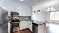 Located at 333 Central Avenue, unit 413 is a well-appointed single-floor residence that combines modern design with functional living. The kitchen is a standout feature, showcasing crisp white shaker-style cabinetry that extends to the ceiling for generous storage capacity. Stainless steel appliances, including a range, microwave, and dishwasher, lend a contemporary and cohesive look throughout the space. A striking mosaic tile backsplash in neutral tones of gray and brown adds texture and visual interest, complementing the dark quartz countertops that provide ample prep and workspace. A kitchen island with an integrated sink and additional countertop space serves as a natural focal point, ideal for casual dining or entertaining. Two sleek metallic pendant lights hang above the island, providing both task lighting and a refined decorative touch. Rich, dark hardwood flooring runs throughout, grounding the overall aesthetic and providing a warm contrast to the bright white cabinetry and light walls. The open layout encourages a seamless flow between the kitchen and the adjoining living areas, making the space feel open and inviting. Clean lines, quality finishes, and thoughtful design details come together to create a modern, polished environment.
