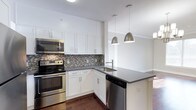 Located at 333 Central Avenue, unit 413 is a well-appointed single-floor residence that combines modern design with functional living. The kitchen is a standout feature, showcasing crisp white shaker-style cabinetry that extends to the ceiling for generous storage capacity. Stainless steel appliances, including a range, microwave, and dishwasher, lend a contemporary and cohesive look throughout the space. A striking mosaic tile backsplash in neutral tones of gray and brown adds texture and visual interest, complementing the dark quartz countertops that provide ample prep and workspace. A kitchen island with an integrated sink and additional countertop space serves as a natural focal point, ideal for casual dining or entertaining. Two sleek metallic pendant lights hang above the island, providing both task lighting and a refined decorative touch. Rich, dark hardwood flooring runs throughout, grounding the overall aesthetic and providing a warm contrast to the bright white cabinetry and light walls. The open layout encourages a seamless flow between the kitchen and the adjoining living areas, making the space feel open and inviting. Clean lines, quality finishes, and thoughtful design details come together to create a modern, polished environment.