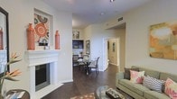 This single-story residence offers an open and well-connected layout that seamlessly integrates the living, dining, and circulation areas into a cohesive living environment. The living room serves as the focal point of the space, anchored by a clean white fireplace with a decorative mantle, above which a vibrant piece of wall art adds a bold pop of color and personality. Rich, dark hardwood flooring flows throughout, providing a warm and grounding contrast to the soft, neutral-toned walls. A glass-topped coffee table and a sage green sofa with accent pillows contribute to the room's comfortable and stylish atmosphere. Adjacent to the living area, a compact dining space accommodates a round table with seating for several guests, positioned near a built-in shelving unit that offers both storage and display opportunities. A framed art piece on the nearby wall adds visual interest to the dining nook. A hallway visible from the main living area leads to additional rooms, suggesting a thoughtful and functional floor plan. Recessed lighting and ceiling vents indicate modern climate control and lighting infrastructure throughout the space.