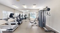 Located at 234 Mallard Point Dr in Coventry Township, OH, this fitness center offers a well-organized and fully equipped exercise space designed to support a comprehensive workout routine. The room features durable speckled flooring throughout, providing a practical and easy-to-maintain surface suited for high-traffic use. Bright flush-mount fluorescent lighting illuminates the space evenly, complemented by natural light streaming in through multiple windows fitted with white slatted blinds. Views of an adjacent outdoor pool are visible through the windows, adding a pleasant ambiance to the workout environment. A large wall-mounted mirror spans a significant portion of one wall, visually expanding the space and allowing users to monitor their form during exercise. The cardio section features multiple True-brand treadmills and an elliptical machine, while the strength training area includes a True multi-station cable machine and a rack of graduated dumbbells. A wall-mounted flat-screen television provides entertainment options, and a wall fan offers additional airflow. Small shelving units and a paper towel dispenser add practical convenience. A doorway connects to an adjacent hallway, suggesting access to additional amenities nearby. The overall atmosphere is clean, bright, and functional.