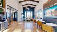 Located in Mount Dora, Florida, this single-floor amenity clubhouse for Yardly Mount Dora offers a thoughtfully designed community space spanning a generous footprint. The main living lounge immediately impresses with soaring vaulted ceilings accented by exposed wood beams, dramatic teal-painted walls, and floor-to-ceiling windows that flood the room with abundant natural light and outdoor views. A sleek linear fireplace set against a floor-to-ceiling stone tile wall anchors the space, complemented by a wall-mounted TV and stylish contemporary furnishings. Light hardwood flooring flows seamlessly throughout. A second living area provides additional gathering space, also bathed in natural light with large multi-pane windows overlooking the community. The fully equipped fitness center is expansive and bright, featuring an extensive array of cardio machines, cable systems, free weights, and functional training equipment, all beneath large ceiling fans and windows offering outdoor views. Motivational signage and mirrored walls enhance the workout environment, along with a hydration station. The dedicated office space offers a sun-drenched work area with pendant lighting, built-in booth seating with lush greenery, and wide views of the surrounding landscape. A stylish kitchen with a marble island, herringbone backsplash, and pendant lighting serves the social areas, alongside a Mount Dora-themed mural wall that adds local character.