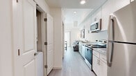 This single-floor residence presents a thoughtfully designed layout that balances functionality and modern aesthetics. The interior is unified by light wood-tone flooring throughout, crisp white walls, and recessed lighting that creates a bright, clean atmosphere.

The galley-style kitchen is well-appointed with stainless steel appliances, including an electric range, over-the-range microwave, dishwasher, and refrigerator. Light wood cabinetry pairs with speckled granite-style countertops, providing ample storage and prep space. A stacked washer and dryer are conveniently located adjacent to the kitchen area.

The kitchen flows directly into the open living area, which benefits from natural light streaming through a large sliding glass door or window at the far end, enhancing the sense of openness.

The unit includes multiple bedrooms, each featuring the same light wood flooring and neutral tones. One bedroom is equipped with a space-saving Murphy bed built into the wall, while another offers a wardrobe, shelving unit, and dresser for practical storage.

The bathroom maintains the unit's clean aesthetic with a light wood vanity, stone-look tile flooring, and simple white panel doors with brushed gold hardware accents.