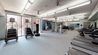 This fitness center in Houston, TX offers a comprehensive workout environment spread across a single level. The space is thoughtfully organized into distinct zones, each designed to support a variety of training styles. Teal and light gray walls provide a clean, energizing aesthetic, complemented by light-toned flooring throughout.

The cardio zone features multiple treadmills and stair climbers positioned near large windows that bring in natural light and views of the surrounding exterior. A decorative striped accent wall adds a dynamic visual element to this section. Ceiling fans and exposed ductwork give the space an industrial yet functional character, while bright linear fluorescent lighting ensures excellent visibility throughout.

The strength training area is generously sized and houses multiple cable and multi-use weight machines arranged with ample space between equipment. A dedicated free weights section includes a full rack of dumbbells in varying sizes, displayed alongside floor-to-ceiling mirrors that enhance the sense of depth. Medicine balls, a punching bag, and a wall-mounted pad round out the functional training area. Mounted flat-screen televisions are positioned throughout for entertainment. The overall layout promotes efficient circulation and supports a full-body training regimen.