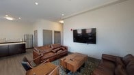 This single-floor space is designed to serve as a versatile communal gathering area, combining lounge, dining, and kitchen functions within an open, well-organized layout. The interior features neutral white walls, wood-look flooring, and recessed lighting throughout, creating a clean and contemporary atmosphere.

The lounge area is furnished with rich brown leather sofas and a sturdy wooden coffee table, anchored by a patterned area rug. A large wall-mounted flat-screen television serves as the focal point of this zone, making it well-suited for group entertainment. Multiple gray doors along one wall lead to additional rooms or storage areas.

Adjacent to the lounge, the dining area accommodates multiple tables paired with yellow upholstered chairs, offering ample seating for group meals or meetings. Tall windows fitted with white blinds line the walls, allowing generous natural light and exterior views into the space.

The kitchen is equipped with dark cabinetry, a stainless steel refrigerator, a dishwasher, and a subway tile backsplash. A prominent curved island with a light-toned granite countertop provides additional workspace and serves as a natural divider between the kitchen and the open living areas. A sliding glass door in the lounge area adds further access to natural light.